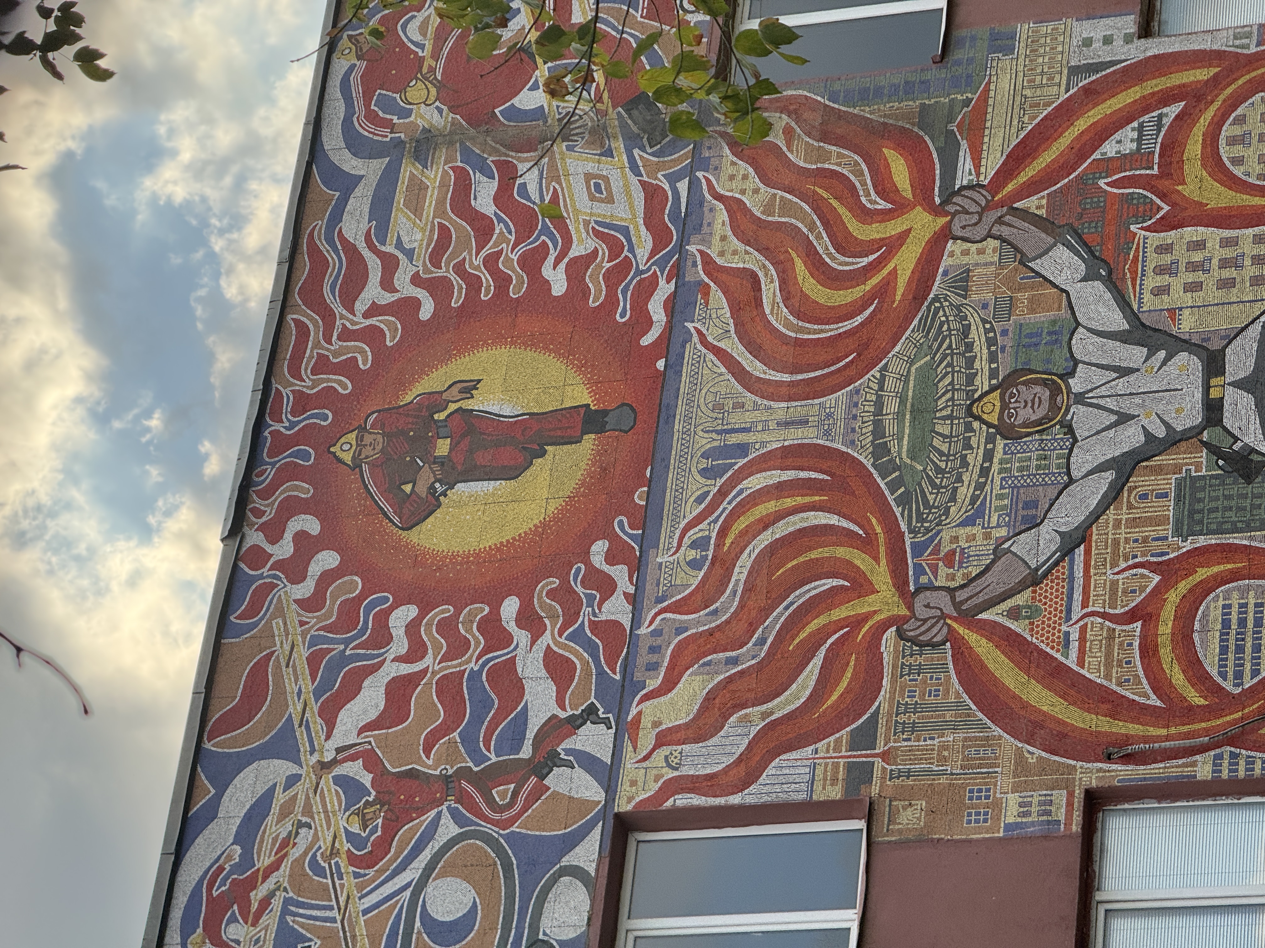 Upper section of the Fire Is in Good Hands mosaic showing red-clad Soviet firefighters running within a sunburst motif in Tbilisi.